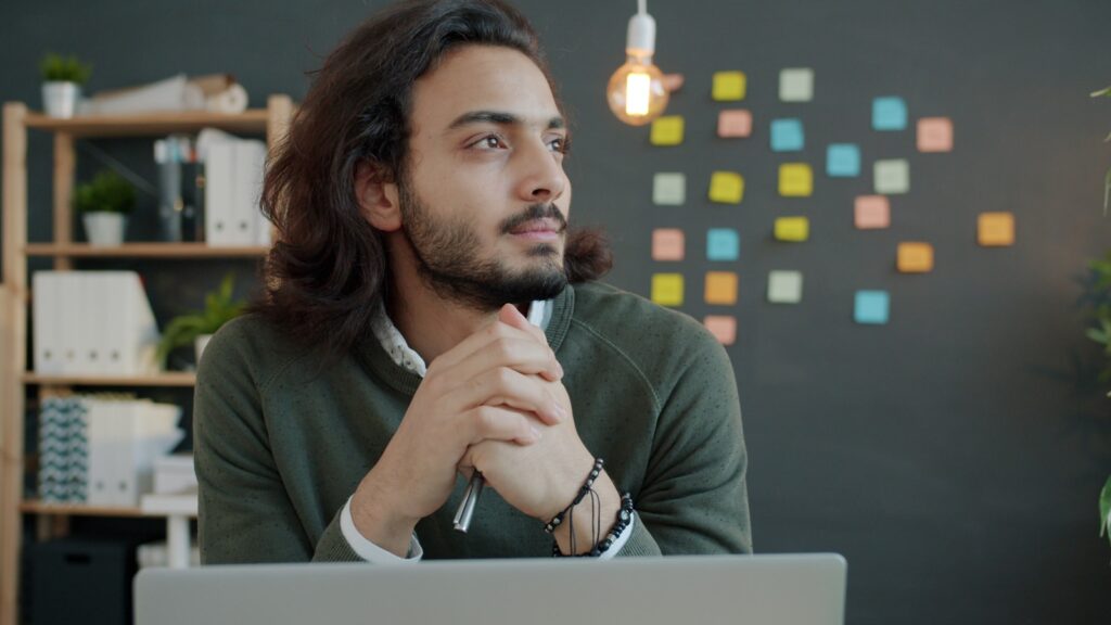 Man with long hair thinking at desk with laptop.